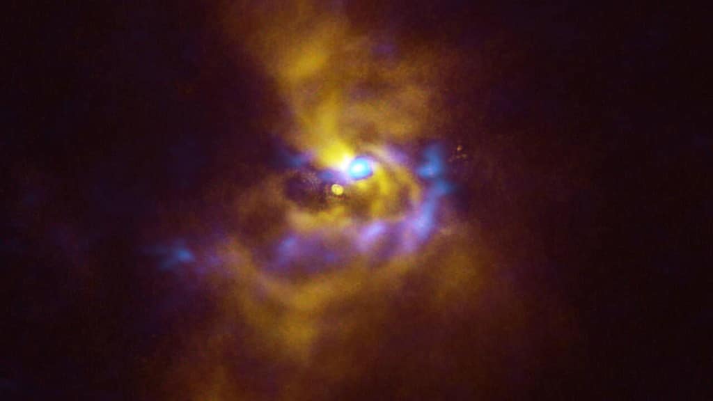 En el centro de esta imagen está la joven estrella V960 Mon, ubicada a más de 5.000 años luz de distancia en la constelación de Monoceros. Rodeando la estrella hay material polvoriento con potencial para formar planetas. Las observaciones obtenidas utilizando el instrumento Spectro-Polarimetric High-contrast Exoplanet REsearch (SPHERE) en el VLT de ESO, representado en amarillo en esta imagen, muestran que el material polvoriento que orbita la joven estrella se está agrupando en una serie de intrincados brazos espirales que se extienden a distancias mayores que todo el Sistema Solar. Mientras tanto, las regiones azules representan datos obtenidos con el Atacama Large Millimeter/submillimeter Array (ALMA), del cual ESO es socio. Los datos de ALMA examinan más profundamente la estructura de los brazos espirales, revelando grandes cúmulos de polvo que podrían contraerse y colapsar para formar planetas gigantes de aproximadamente el tamaño de Júpiter a través de un proceso conocido como "inestabilidad gravitacional".