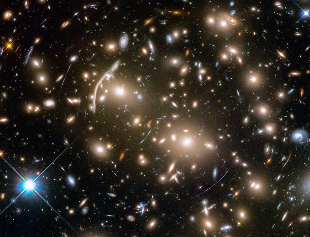 El cúmulo de galaxias Abell 370, ubicado a unos 4 mil millones de años luz de distancia, contiene una asombrosa variedad de varios cientos de galaxias unidas por la atracción mutua de la gravedad. Enredados entre las galaxias hay arcos de luz azul de aspecto misterioso. En realidad, se trata de imágenes distorsionadas de galaxias remotas detrás del cúmulo. Estas galaxias remotas son demasiado débiles para que el Hubble pueda verlas directamente. En cambio, la gravedad del cúmulo actúa como una enorme lente en el espacio que magnifica y amplía las imágenes de las galaxias de fondo como un espejo de feria.